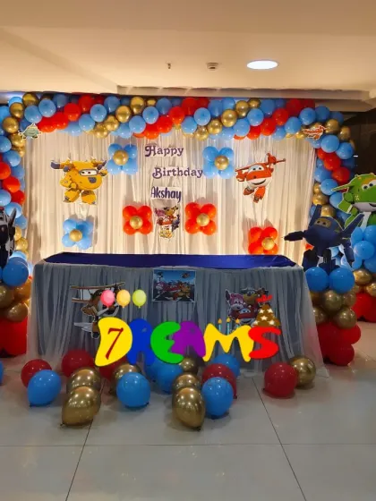 A Super Wings themed birthday party. This setup includes a balloon arch in red, blue, and gold, with character cutouts of the airplanes against a simple white curtain backdrop.