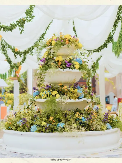 A three-tiered fountain overflowing with a vibrant mix of yellow, blue, and purple flowers, acting as a stunning central decor piece for the Mehendi.