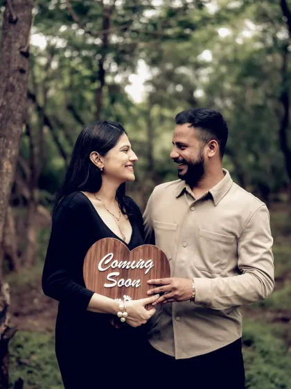 The couple stands together in a forest clearing, holding a "Coming Soon" sign. Their shared smile shows their mutual excitement for the future.
