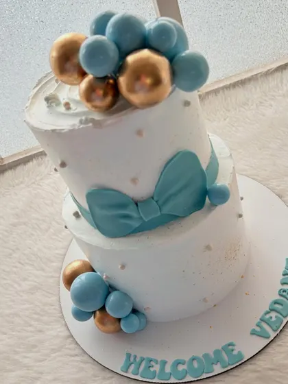 A beautiful two-tier welcome cake for a baby boy. The design is elegant and simple, with a soft blue bow and clusters of blue and gold spheres.
