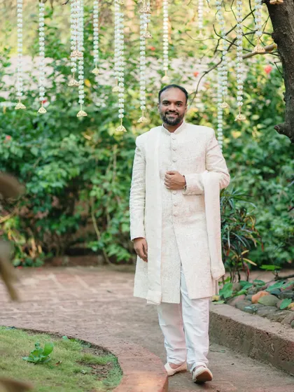 A full-length portrait of the groom in a classic white sherwani. The natural garden setting provides a perfect backdrop for this elegant and timeless shot.