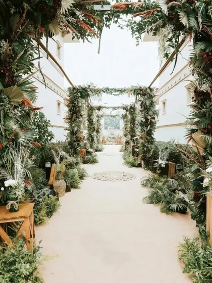 A rustic and lush walkway for a nature-inspired wedding. The path is lined with an abundance of ferns, tropical leaves, and wooden crates, creating an enchanted forest feel.