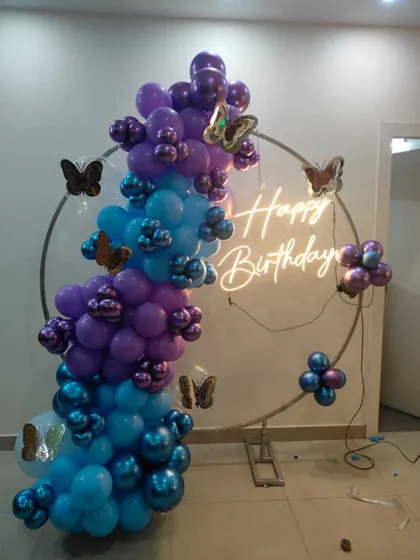 A birthday balloon arrangement on a circular frame. The balloons in shades of purple and blue are accented with butterfly stickers and a neon sign.
