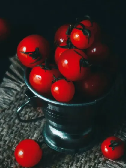 A moody shot of vibrant cherry tomatoes in a small metal bucket on a rustic burlap surface. The dark, dramatic lighting makes the red color pop and highlights the freshness of the produce.