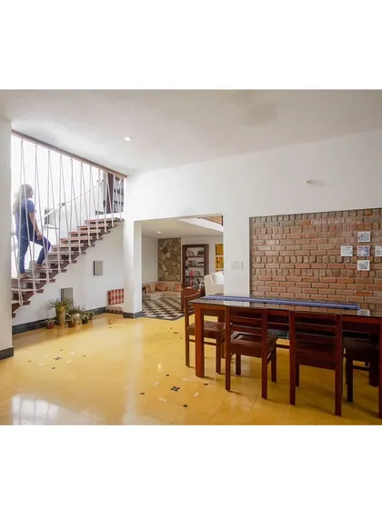 The open-plan layout of 'Malenadu Mane' connects the dining area with the staircase and other living spaces. The use of exposed brick and warm wood tones creates a cohesive and welcoming atmosphere.