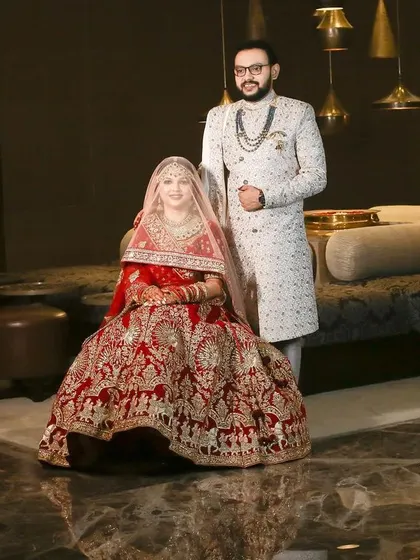 A regal wedding portrait. The bride is seated in her magnificent red lehenga while the groom stands beside her, creating a powerful and elegant image of the couple on their big day.