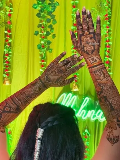 A bride celebrating at her mehendi function, with her beautifully decorated hands raised. The neon "Mehndi" sign in the back adds to the festive atmosphere.