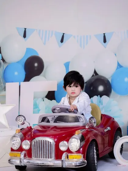 This one-year-old is ready to drive into his second year, sitting in a red toy car with a blue and white balloon backdrop.