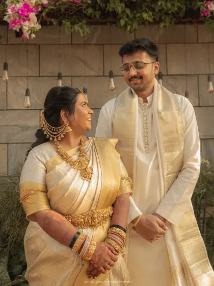 A portrait of a couple in matching white and gold traditional wear. Their comfortable smiles and shared look speak of a deep and happy connection.