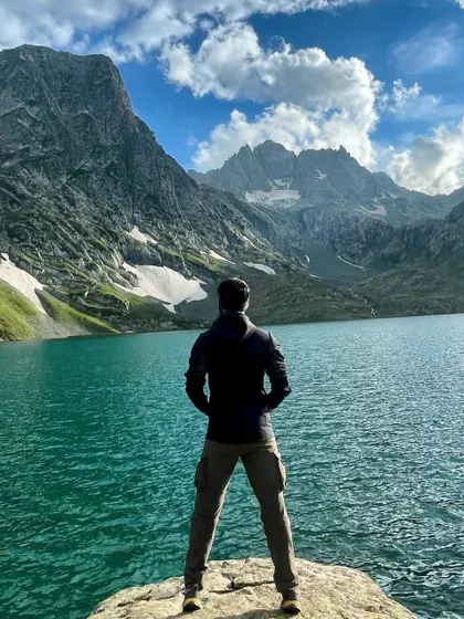 Lost in the vastness of nature. A trekker contemplates the serene, turquoise waters of a lake on the Kashmir Great Lakes trek, a journey through some of the most stunning landscapes on earth.