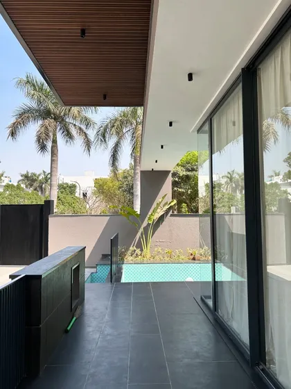 This view from the covered veranda at Gulmohar Farmhouse shows how the pool is integrated directly with the living space. The wooden ceiling and dark floor tiles create a warm, resort-like feel, with views of palm trees framing the scene.