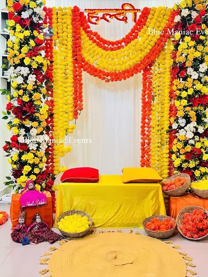 A beautiful Haldi setup with a frame of red, yellow, and white flowers and marigold garlands. The traditional Rajasthani puppets add a touch of folk art to the decor.