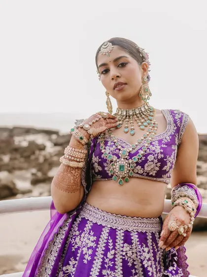 A striking portrait of a bride in a vibrant purple lehenga, posing against a beachside railing, her look confident and modern.