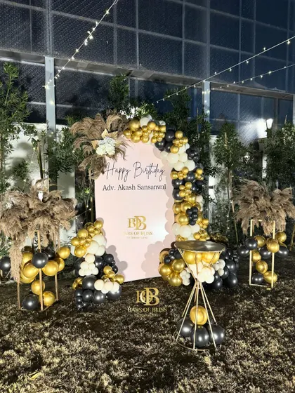 An elegant outdoor birthday party setup perfect for an evening celebration. The decor features a personalized backdrop framed by black, white, and gold balloons, with pampas grass arrangements adding a touch of softness.