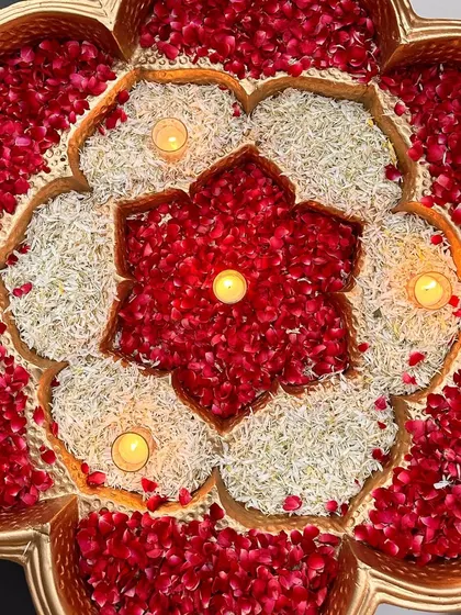 A beautiful floral rangoli made of red and white petals in a brass urli, lit with candles.
