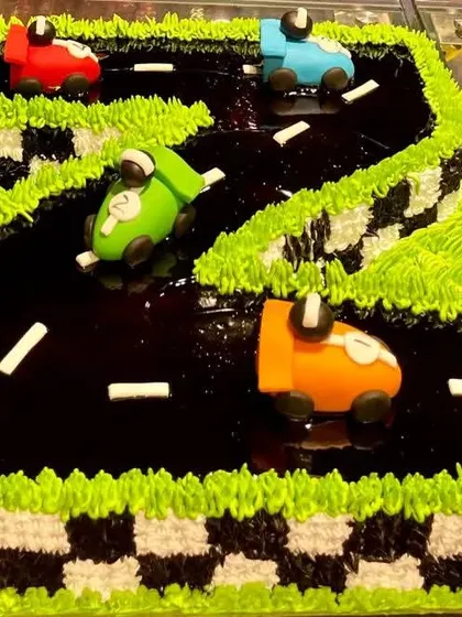 A race car track cake shaped into the number two for a second birthday. The track is made of dark chocolate glaze, with colorful fondant cars racing along it.