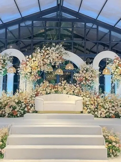 A beautiful glasshouse reception setup with a stage full of white and pastel flowers under elegant arches.