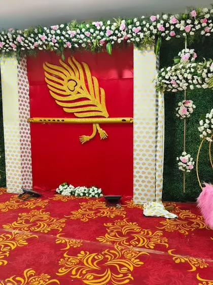 A Krishna-themed backdrop for a naming ceremony. The setup includes a red and green wall, a large golden peacock feather and flute cutout, and a cradle decorated with pink fur.