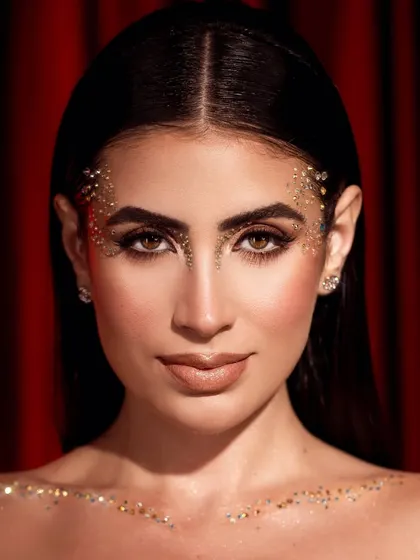 A creative beauty shot with scattered glitter on the face and collarbones, set against a rich red curtain for a theatrical feel.