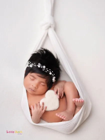 A creative and artistic shot of a baby sleeping in a white fabric sling, holding a small heart. This pose is beautiful, simple, and elegant.