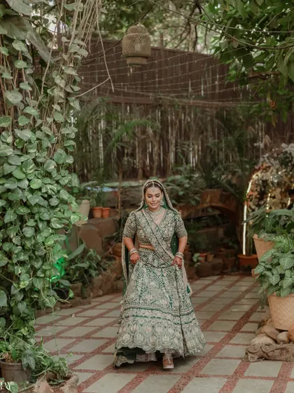 The bride walks through a lush, green pathway, her beautiful green lehenga perfectly complementing the natural surroundings.