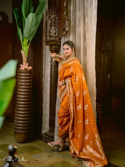 A regal portrait of a bride in a stunning orange Nauvari saree, leaning against a traditional carved wooden pillar.