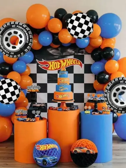 A Hot Wheels themed birthday party setup with a checkered backdrop, orange and blue balloons, and themed pedestals for the cake and desserts.