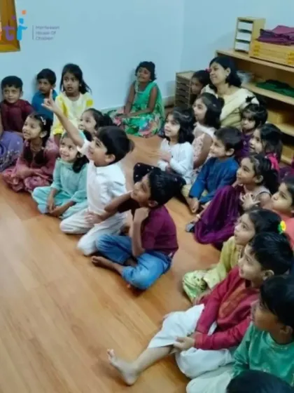 Children sit on the floor, completely engrossed in a storytelling session. One child raises his hand, eager to ask a question, showing his active engagement in the learning process.