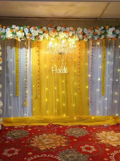 A simple yet elegant Haldi backdrop with a floral arch made of white and orange flowers. A chandelier and a 'Haldi' neon sign add a touch of glamour.