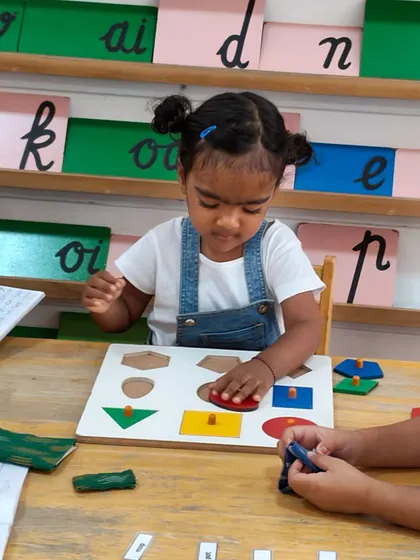A young child works with the geometric shape puzzle. This activity not only teaches the names of shapes but also develops hand-eye coordination and problem-solving skills as she fits each piece into its correct place.