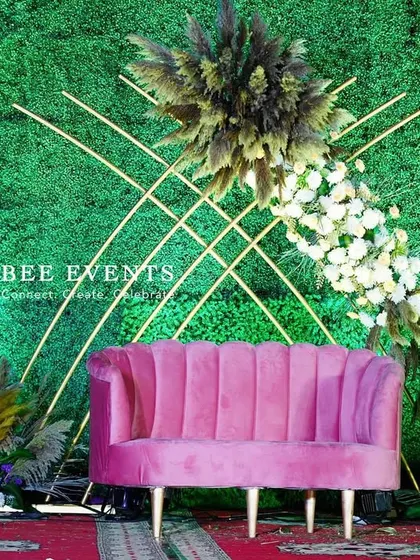A close-up of our unique reception stage with a green backdrop. The design features a striking diagonal floral arrangement, geometric gold lines, and a chic pink sofa, perfect for a modern and artistic couple.