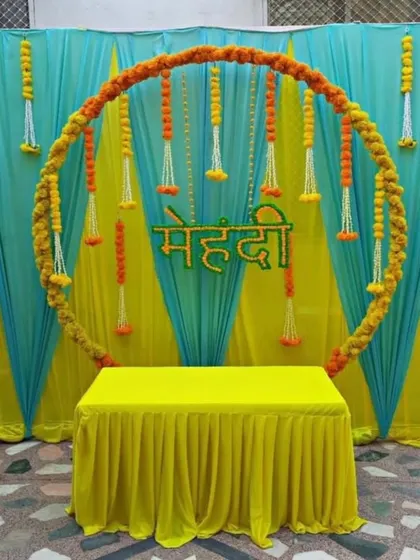A clean and modern Mehndi setup with a circular marigold ring as the centerpiece. The backdrop uses a combination of turquoise and yellow drapes with hanging floral strings, creating a fresh and bright look.