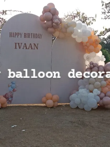 A large outdoor backdrop for a birthday party on a lawn. The setup includes custom arched panels and a balloon garland in a muted, elegant color palette.