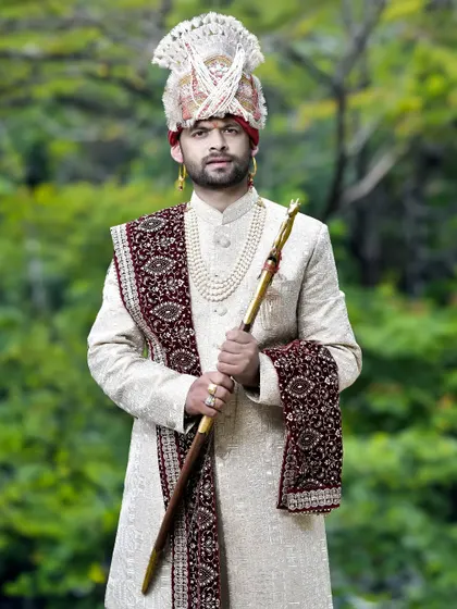 A regal groom's portrait in a cream sherwani and elaborate turban, holding a ceremonial staff. The outdoor setting provides a beautiful, natural backdrop.