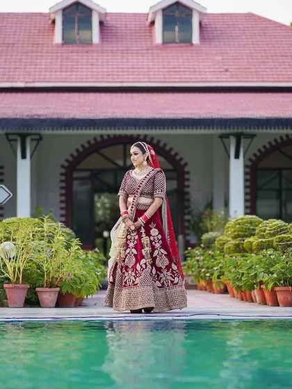 A grand bridal portrait by the pool. The bride stands in her red lehenga against the backdrop of a beautiful estate, creating a stunning and majestic image.