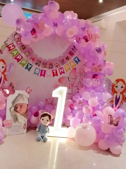 A fairy-themed first birthday with a personalized touch. The setup includes a pink and purple balloon arch, a "Happy Birthday" banner, fairy cutouts, a light-up number '1', and a poster with the baby's photos.