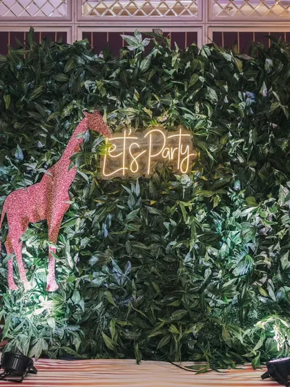 A fun photo booth for the "Wild One" party, featuring a wall of green leaves, a sparkling pink giraffe, and a "Let's Party" neon sign.