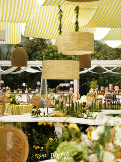 A view across the custom-designed, serpentine-shaped central table. The hanging lampshades and beehives create a stunning visual canopy, while the table itself is filled with lush, garden-like floral arrangements.