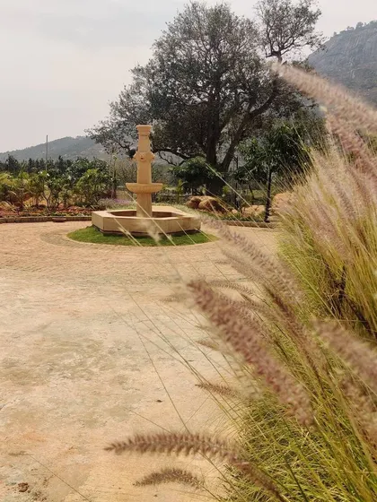 A stone fountain serves as a tranquil focal point in the garden of the Nandi Hills weekend home. The surrounding landscape features a fruit orchard with pomegranate, chikoo, and mango trees.