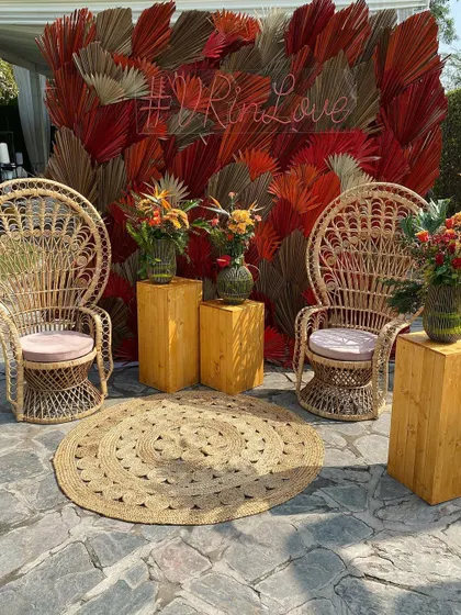A perfect photo spot for a boho-chic event. This seating nook features iconic peacock chairs set against a backdrop of dried red palm leaves, combining natural textures with a pop of color.