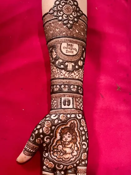 A close-up of a baby shower mehandi featuring a portrait of baby Krishna on the palm. The design extends up the arm with motifs like footprints and a 'Baby Coming Soon' message.
