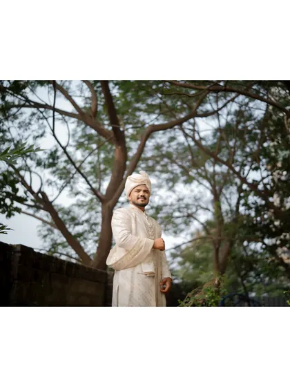 A wide shot of the groom standing under a canopy of trees, looking dapper in his white sherwani and turban.