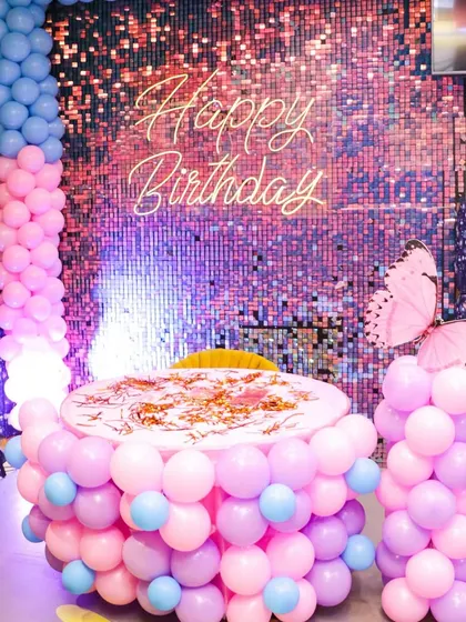 A beautiful butterfly-themed birthday setup. The cake table is wrapped in pastel pink and blue balloons, complemented by a shimmering backdrop and large butterfly accents for a magical feel.