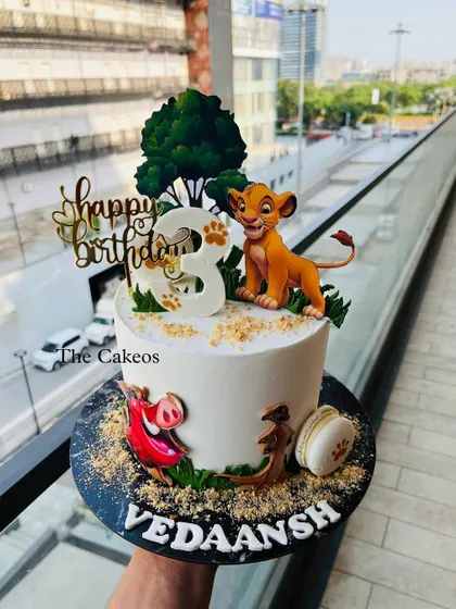 A classic Lion King themed cake for a third birthday. It features a young Simba topper alongside Timon and Pumbaa, set against a tree and a sandy base, perfect for a Disney fan.