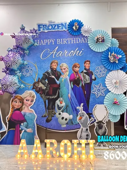 A complete Frozen theme setup with a large backdrop featuring all the characters. The decor is accented with paper fan snowflakes and character standees.