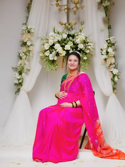 A stunning studio portrait of a mother-to-be in a vibrant pink saree, seated against an elegant white floral backdrop. This setup combines traditional attire with a modern, decorative aesthetic.