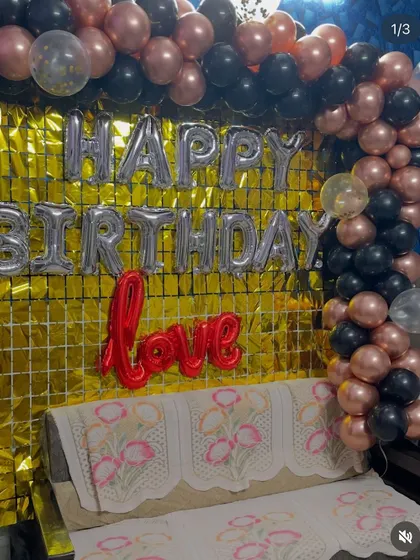 A birthday decoration with a gold shimmer curtain and a black and rose gold balloon arch.