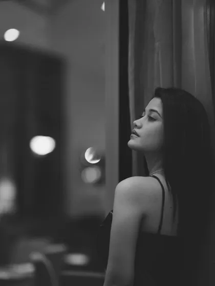 A soft, moody black and white portrait of a woman looking out a window. The reflection and out-of-focus background lights add a layer of depth and contemplation to the scene.
