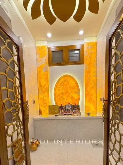 A view through the beautiful floral jali doors into the radiant puja room. The backlit onyx panel and lotus ceiling design are perfectly framed by the doorway.