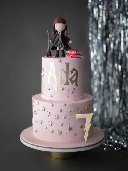 A magical Harry Potter inspired cake for a 7th birthday. This pink and gold two-tiered cake features a Hermione-like figure and the iconic lightning bolt.